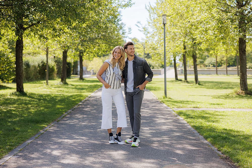 Ein lächelndes Paar spaziert an einem sonnigen Tag einen sonnenbeschienenen Parkweg entlang, umgeben von grünen Bäumen.